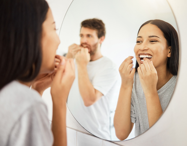 Mirror, dental floss and teeth healthcare couple cleaning mouth for healthy gums, smile and fresh breath. Man and woman in bathroom flossing morning during routine for dental hygiene in the morning Couple flossing looking in bathroom mirror